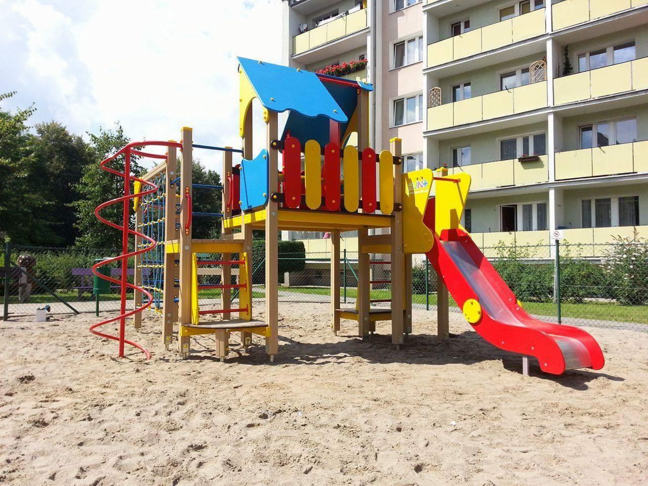 Kolorowy plac zabaw z czerwoną zjeżdżalnią i spiralną drabinką na piaszczystym podłożu, na tle bloku mieszkalnego z balkonami.