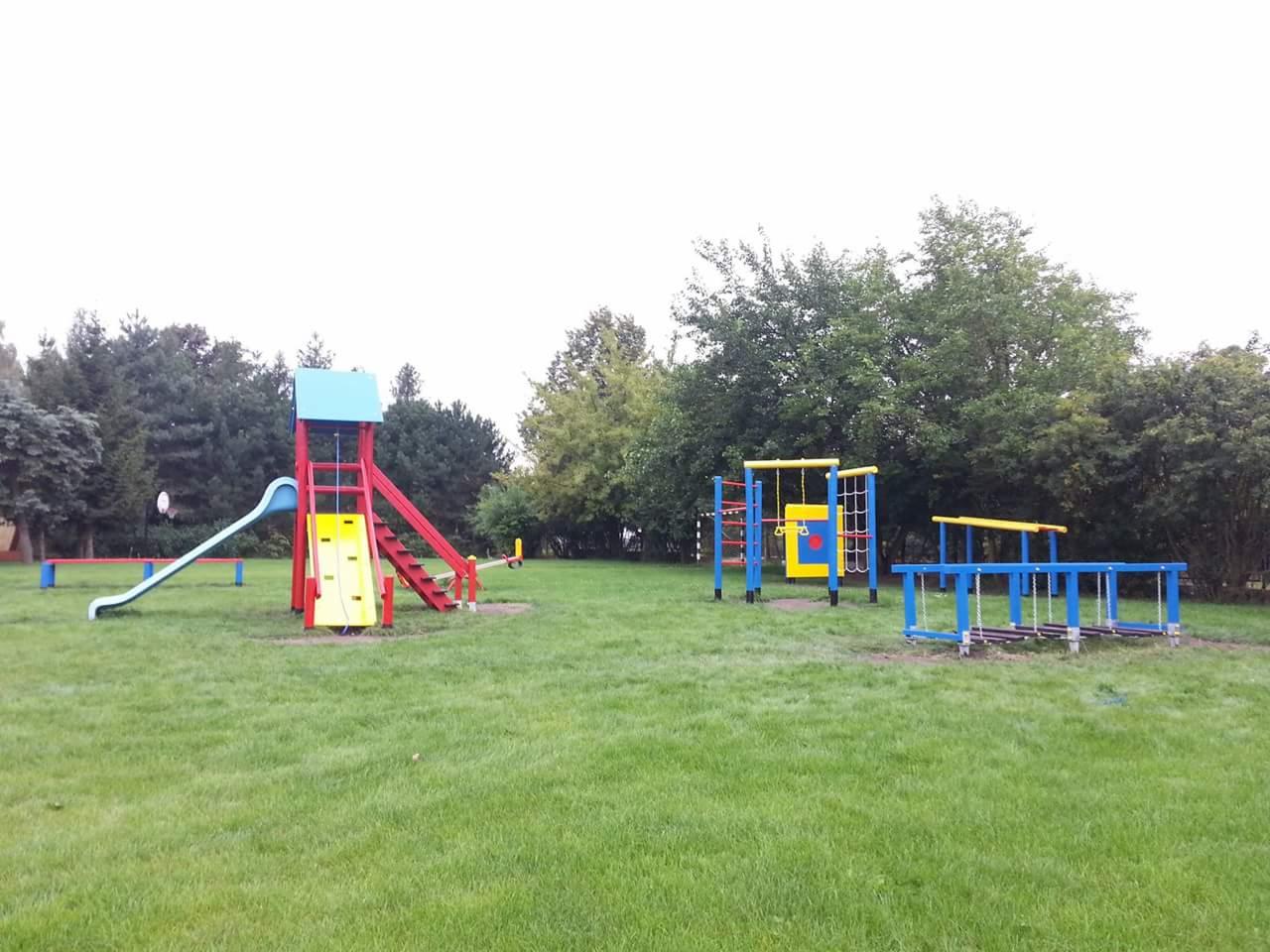Kolorowy plac zabaw z wieżą ze zjeżdżalnią, drabinkami i huśtawkami linowymi na trawiastym terenie, otoczony drzewami.