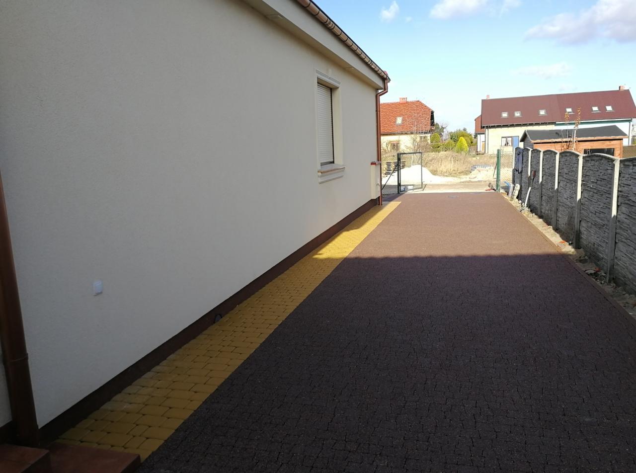 Brązowa nawierzchnia z kostki brukowej, obrzeże z żółtej kostki, przylegająca do ściany budynku z oknem i szarym ogrodzeniem panelowym w tle.