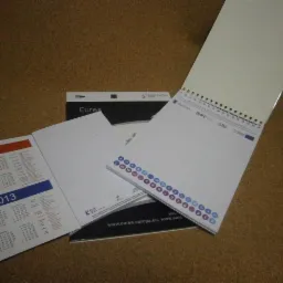 Rozłożony kalendarz książkowy z datami 2012 i 2013 obok kalendarza biurkowego na spirali z logo EURES, leżące na korkowej powierzchni.