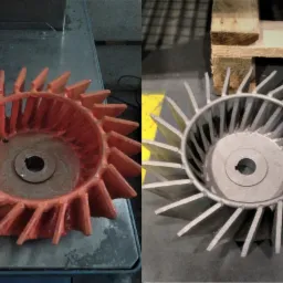 Porównanie dwóch metalowych wirników wentylatorów, jeden pomalowany na pomarańczowo po lewej stronie, drugi surowy, srebrny po prawej stronie, leżący na palecie.