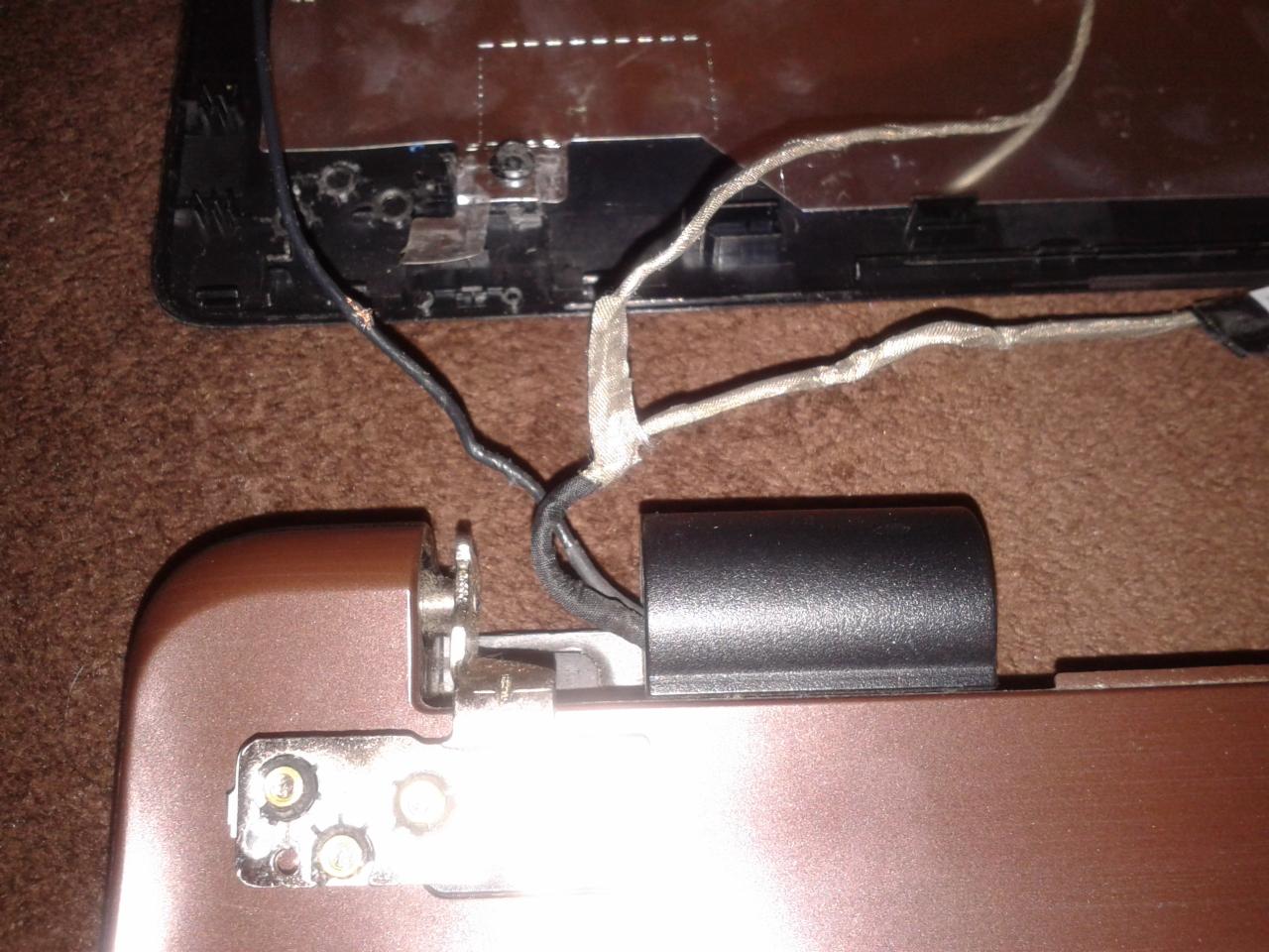 Rozłożony laptop z widocznymi przewodami i elementami mocującymi ekran, brązowa obudowa i fragment ekranu na brązowym tle.