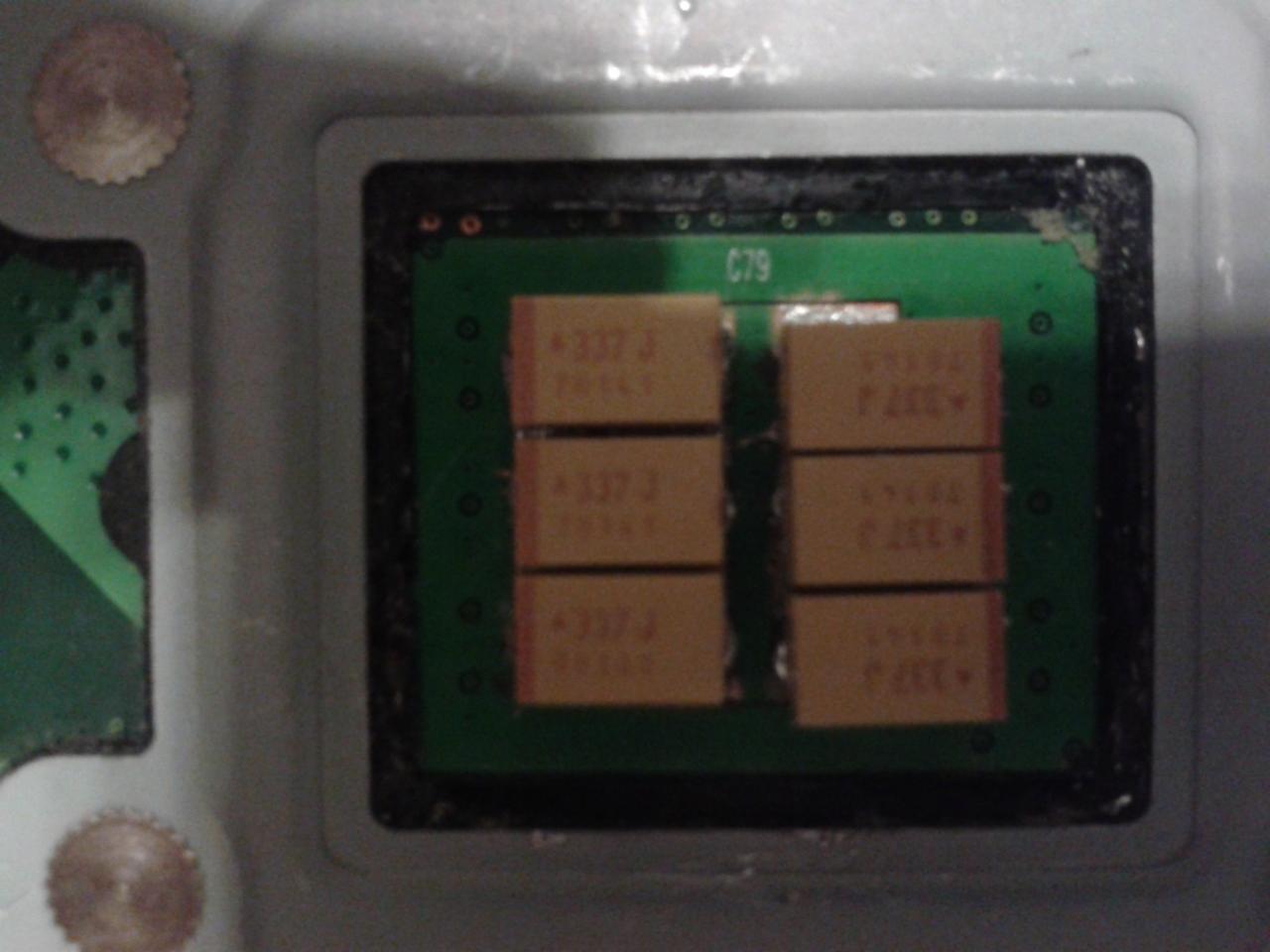 Zbliżenie na płytkę drukowaną z układem kondensatorów oznaczonych +337J, umieszczonych w dwóch kolumnach. Widoczny napis C79 na zielonej powierzchni płytki. Płytka otoczona czarną ramką, a całość...