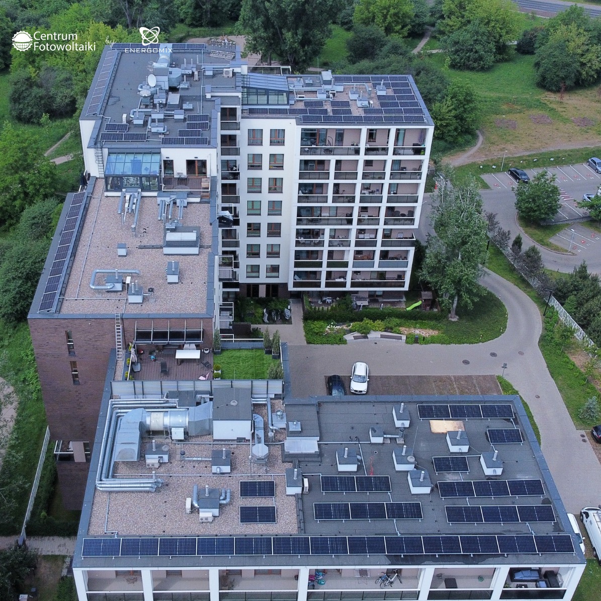 Instalacja fotowoltaiczna na dachach budynków wielorodzinnych, widok z góry, panele rozmieszczone na różnych poziomach, zieleń w tle, logo firmy w lewym górnym rogu.