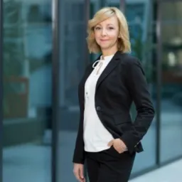 Portret uśmiechniętej kobiety w czarnym garniturze i białej bluzce, stojącej na tle przeszklonej fasady budynku z widocznymi roślinami wewnątrz.