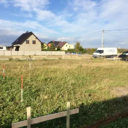 Wytyczony teren pod budowę z ustawionym statywem niwelacyjnym, palikami oznaczającymi granice i drewnianymi elementami ogrodzenia, widoczne budynki w tle oraz samochody