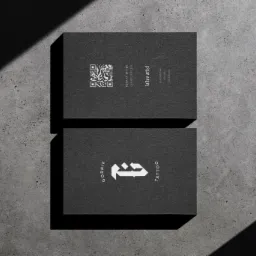 Dwie czarne wizytówki leżące na szarym, betonowym tle. Górna wizytówka zawiera kod QR i dane kontaktowe, a dolna logo firmy z napisem GORNIK TATTOO. Na wizytówki pada cień.