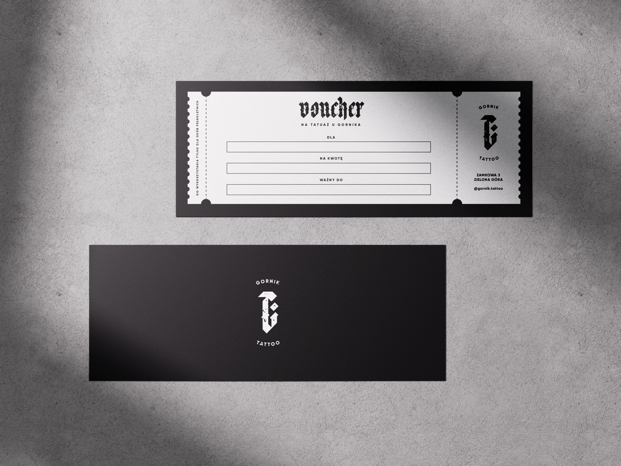 Dwa vouchery prezentowe z logo studia tatuażu Górnik Tattoo, jeden biały z czarnymi napisami, drugi czarny z białym logo, leżące na szarym, teksturowanym tle.