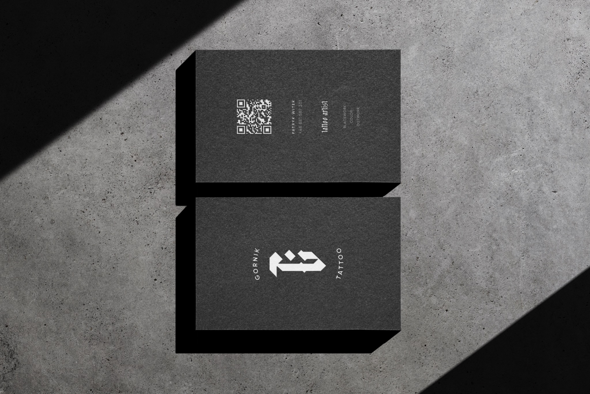 Dwie czarne wizytówki leżące na szarym, betonowym tle. Górna wizytówka zawiera kod QR i dane kontaktowe, a dolna logo firmy z napisem GORNIK TATTOO. Na wizytówki pada cień.