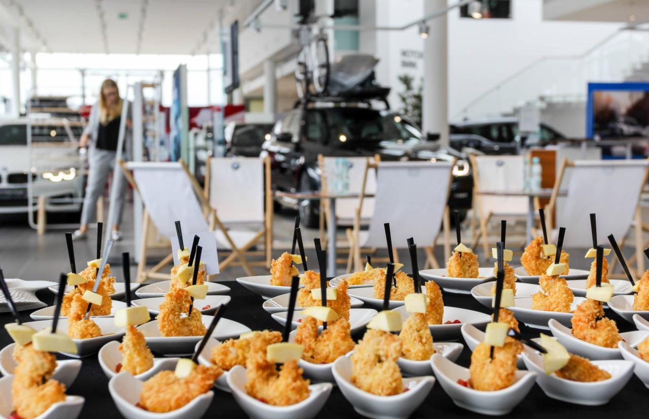 Ustawione na czarnym stole małe białe miseczki z krewetkami w tempurze i kostką sera, czarne patyczki, w tle salon samochodowy z samochodami BMW i leżakami.