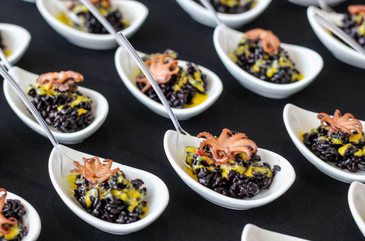 Elegancko podane porcje czarnego ryżu z sosem i małą ośmiornicą na białych łyżkach, ułożone na czarnym tle.