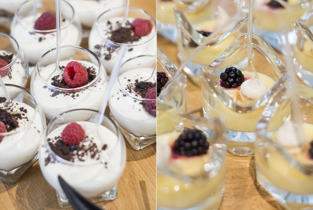 Desery w małych szklankach: biały krem z malinami i posypką czekoladową oraz żółty krem z jeżynami i kuleczką bitej śmietany, podane na drewnianym stole.