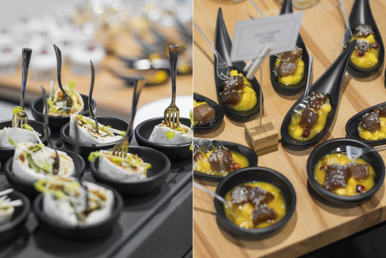 Dwa rodzaje przystawek w czarnych naczyniach: po lewej roladki z białego ciasta z zielonym farszem i widelczykami, po prawej kawałki ryby w żółtym sosie z posypką na drewnianej desce.