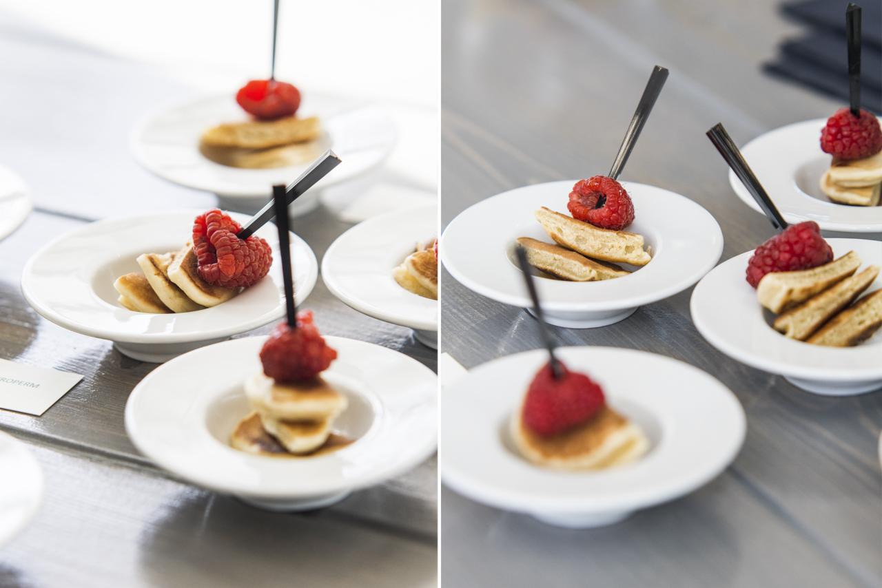 Elegancko podane mini-pancakes z malinami na białych, designerskich talerzykach, udekorowane czarnymi szpikulcami, na gładkim, szarym stole.