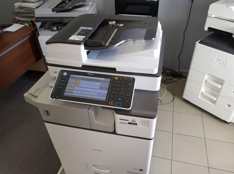 Biała kserokopiarka Ricoh z panelem dotykowym, ustawiona w biurze na tle innych urządzeń, widoczna naklejka serwisowa.