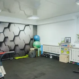 Wnętrze siłowni z tapetą w heksagony, sprzętem do ćwiczeń, piłkami gimnastycznymi i regałem z akcesoriami fitness.