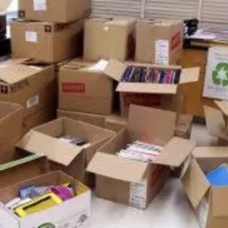 Stos kartonowych pudeł z zawartością, przygotowanych do transportu, z naklejką recyklingu w tle.