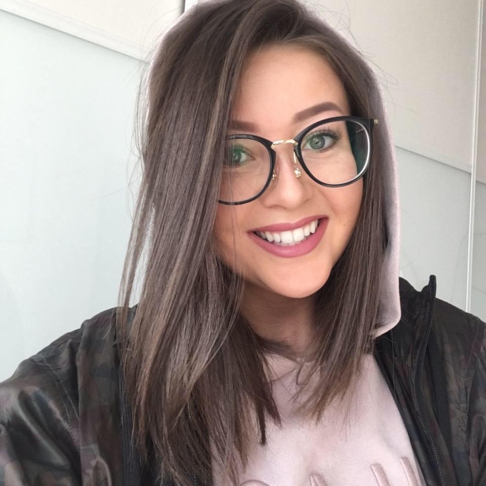 Uśmiechnięta brunetka w okularach i kurtce moro, z delikatnym makijażem, pozuje na tle białej ściany. Widoczne zielone oczy i proste, brązowe włosy.