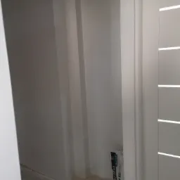 Pomalowane na biało ściany w narożniku pokoju, widoczna torba z gładzią MP Finish przy ścianie oraz fragment białych drzwi z poziomymi panelami.