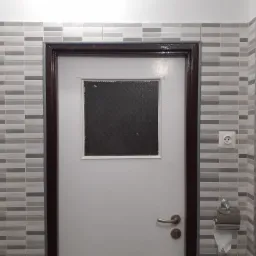 Ściana łazienki wyłożona szarymi, podłużnymi płytkami, białe drzwi z ciemną ramą i małym okienkiem z matowego szkła, widoczne gniazdka elektryczne i uchwyt na papier toaletowy.