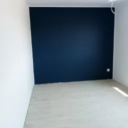Pokój po remoncie: ściana pomalowana na granatowo, sąsiadująca z białą ścianą i jasną podłogą z paneli, widoczne gniazdko elektryczne.