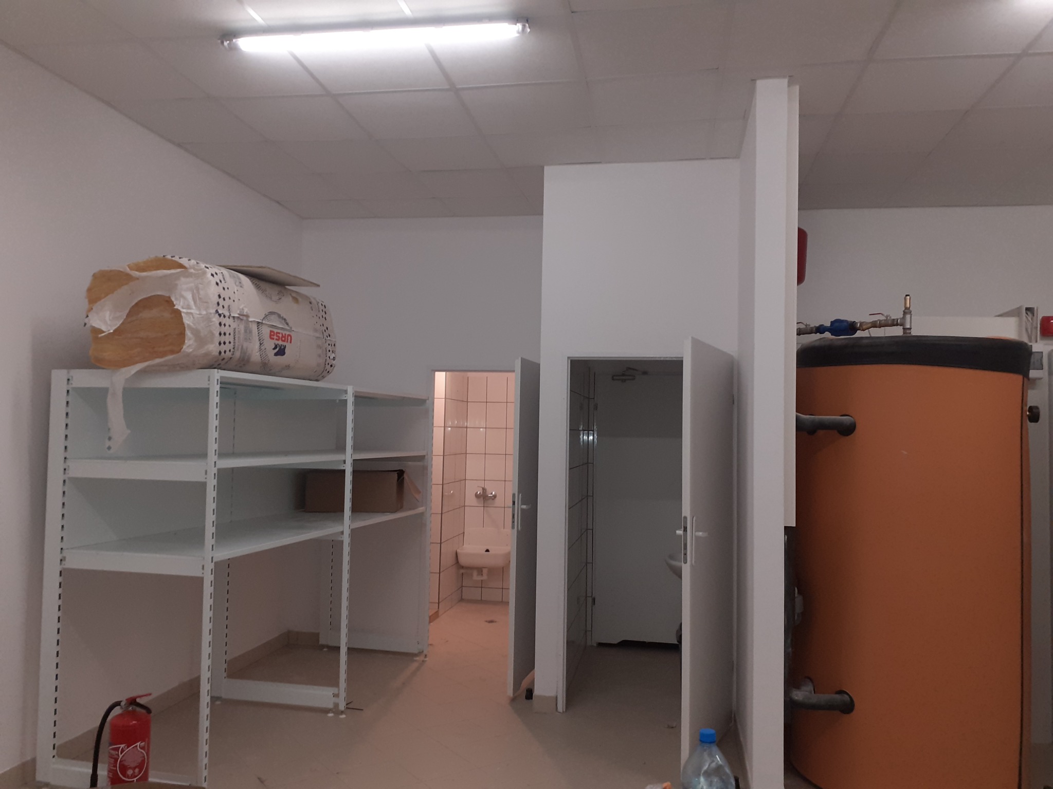 Wnętrze pomieszczenia gospodarczego w trakcie prac wykończeniowych: regał z wełną mineralną, bojler, widoczne dwie łazienki w stanie surowym.