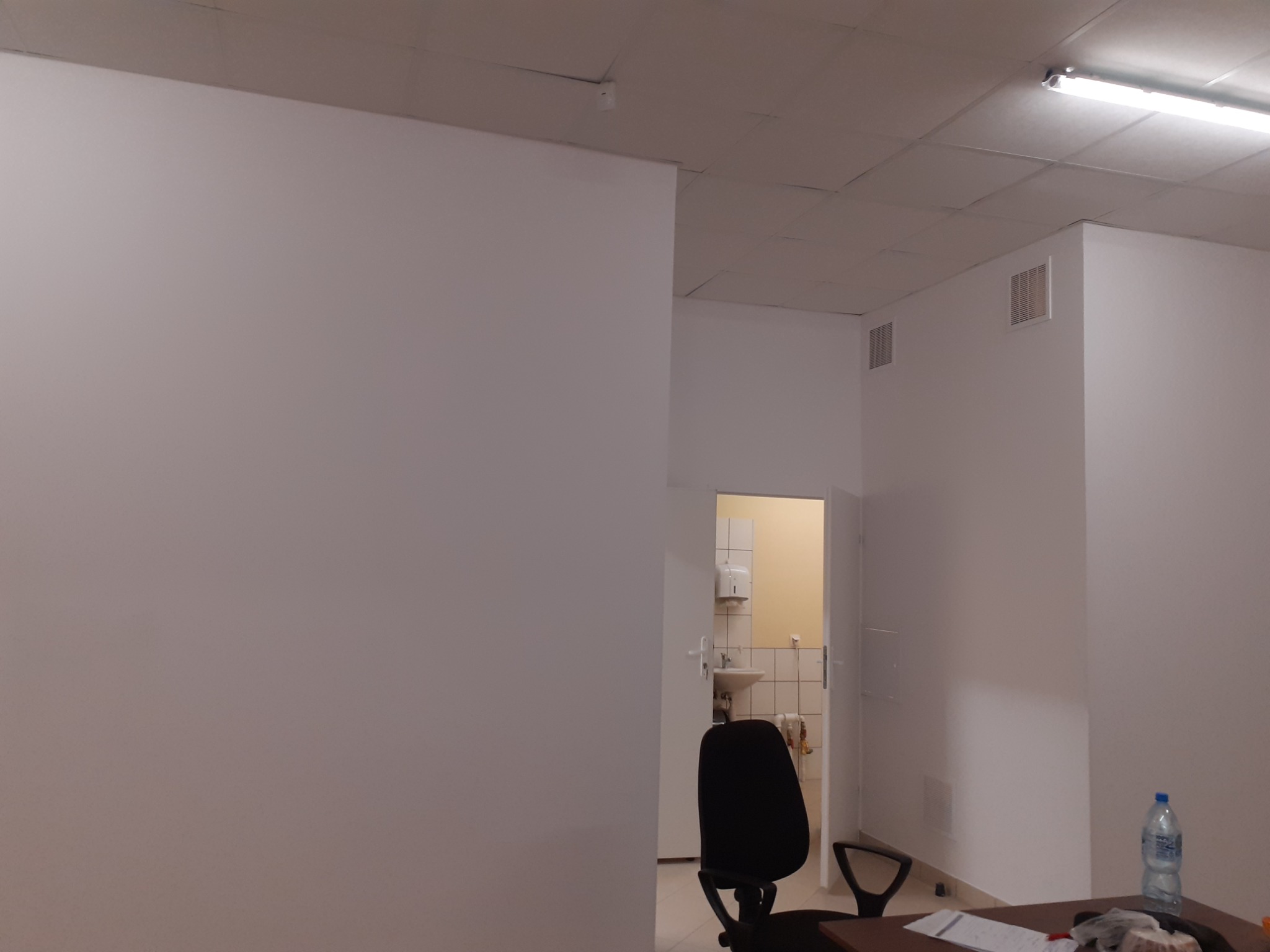 Wnętrze biura po odświeżeniu: białe ściany, sufit z kasetonami, widoczne kratki wentylacyjne, częściowo widoczne biurko z krzesłem i butelką wody, otwarte drzwi do łazienki w tle.