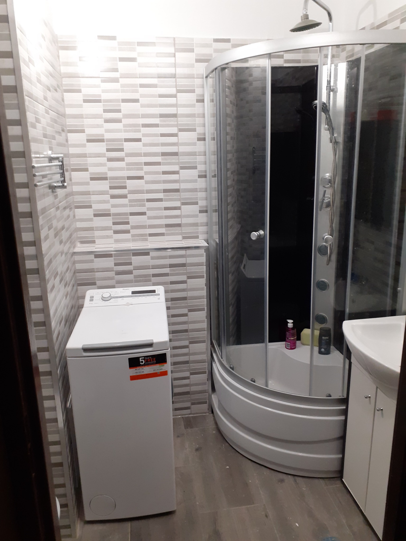 Ukończona łazienka z kabiną prysznicową, umywalką z szafką i pralką, wyłożona płytkami w odcieniach szarości.