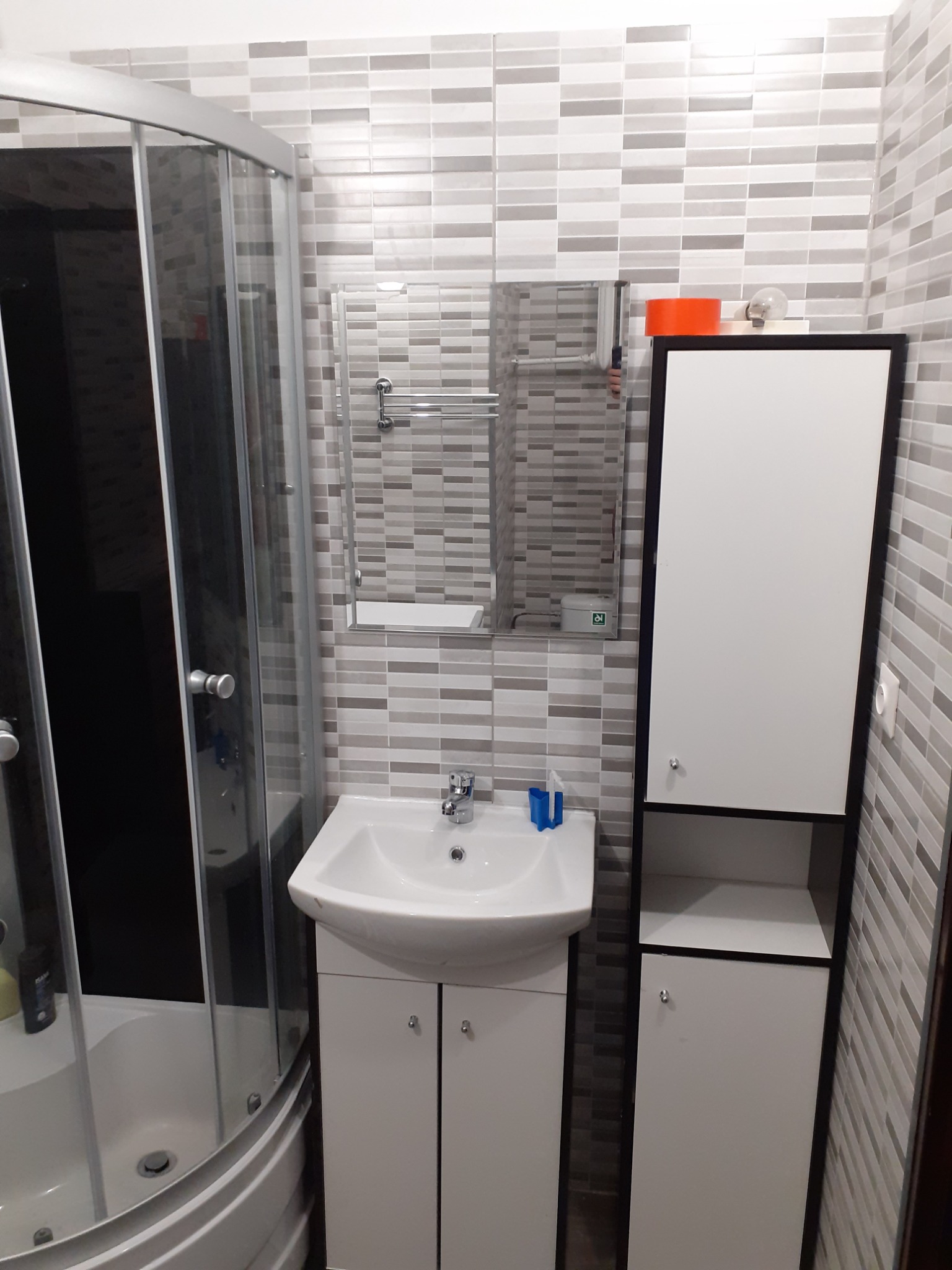 Ukończona łazienka z kabiną prysznicową, umywalką z szafką i wysoką szafką, wykończona płytkami w odcieniach szarości.