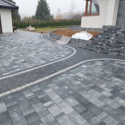 JAKUB GŁĄB PERFEKT-BRUK - Nowoczesna posesja z szarej kostki brukowej ułożonej we wzór, z elementami kostki granitowej i schodami z kamienia. Widok na budynek i zadbany ogród w tle.