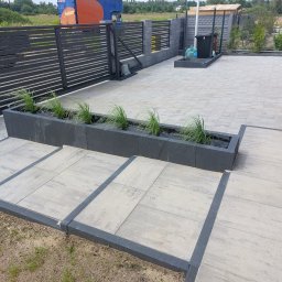 JAKUB GŁĄB PERFEKT-BRUK - Nowoczesna aranżacja przestrzeni: jasnoszara kostka brukowa z ciemnymi obrzeżami, podwyższona rabata z zielenią i automatyczna brama w tle. Minimalistyczny design.