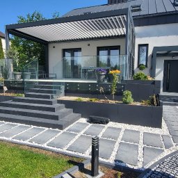 JAKUB GŁĄB PERFEKT-BRUK - Nowoczesny ogród z tarasem i pergolą. Szara kostka brukowa ułożona w ścieżkę, białe kamienie ozdobne. Minimalistyczny design z elementami zieleni i szklanymi balustradami.