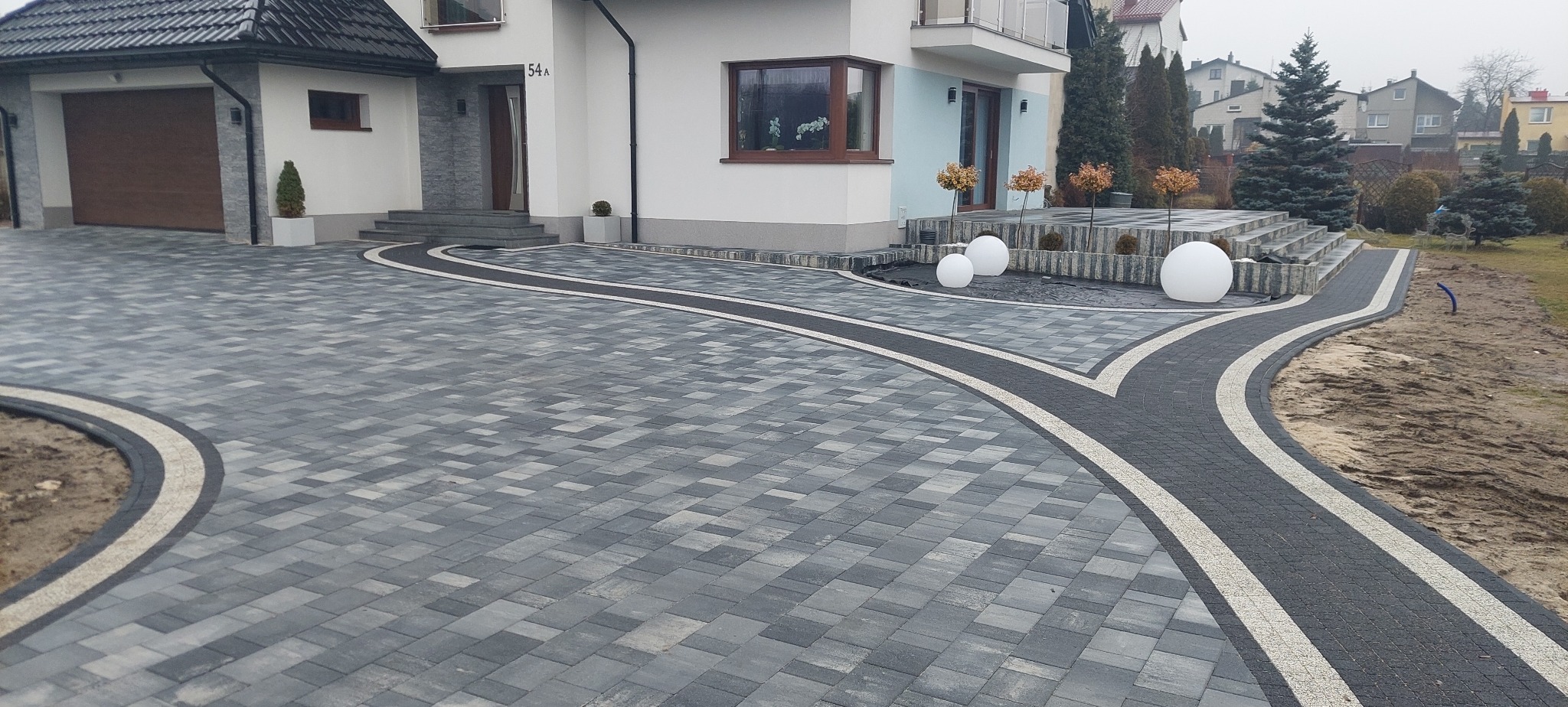 Elegancka posesja z szarej kostki brukowej, z białymi obrzeżami i dekoracyjnymi kulami. Widok na dom z garażem i schodami.