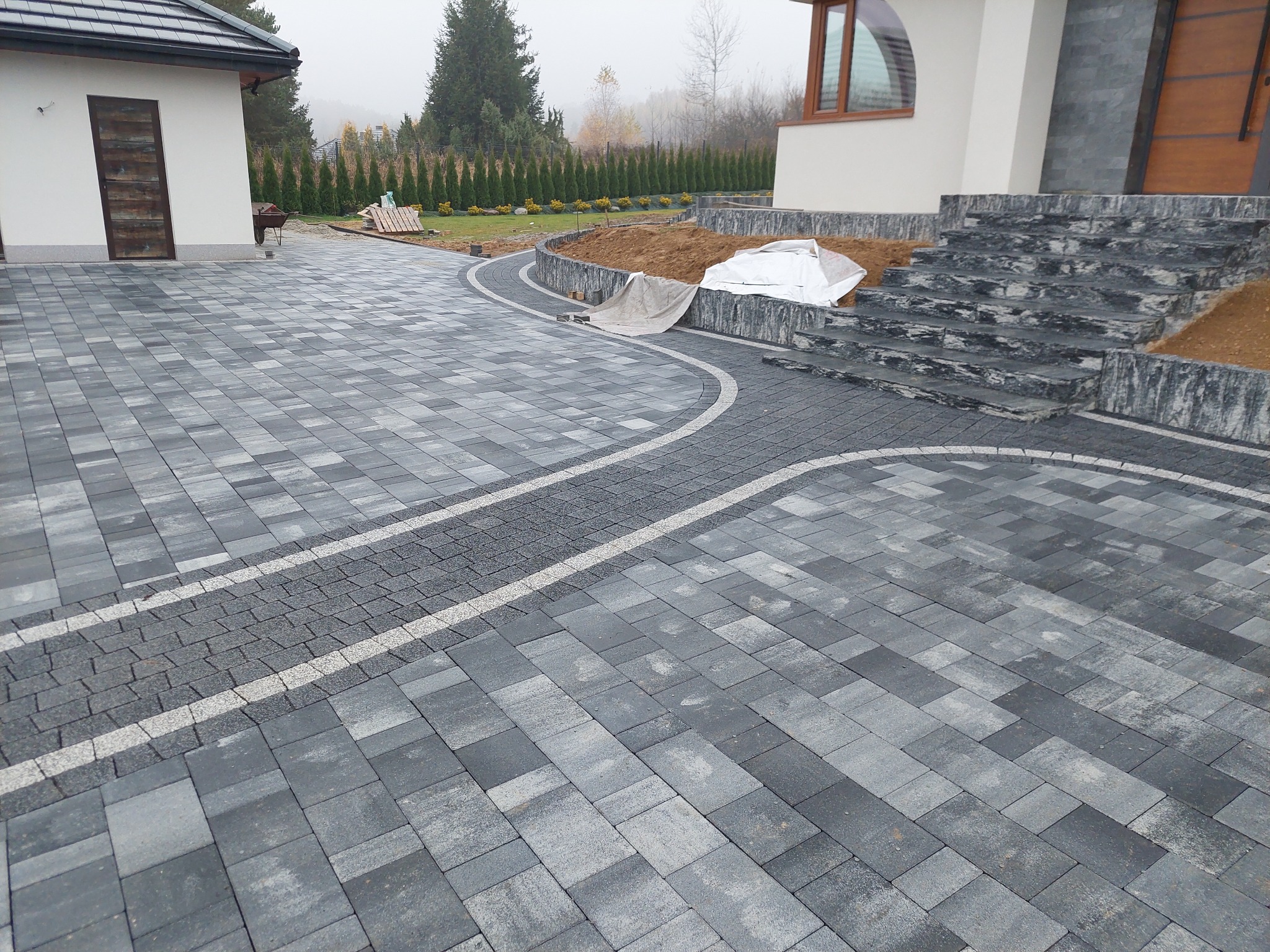 Nowoczesna posesja z szarej kostki brukowej ułożonej we wzór, z elementami kostki granitowej i schodami z kamienia. Widok na budynek i zadbany ogród w tle.