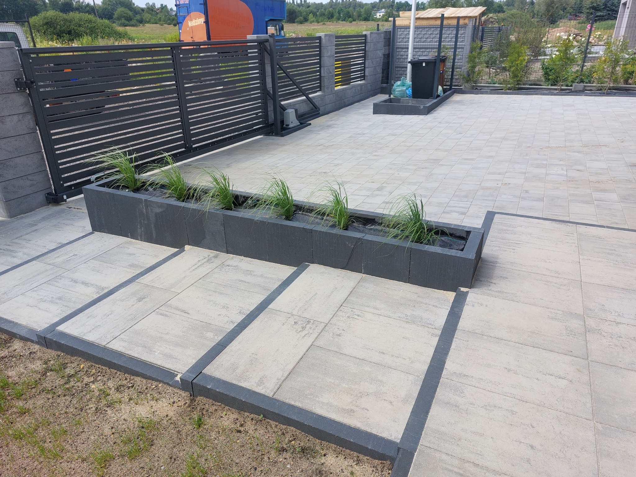 Nowoczesna aranżacja przestrzeni: jasnoszara kostka brukowa z ciemnymi obrzeżami, podwyższona rabata z zielenią i automatyczna brama w tle. Minimalistyczny design.