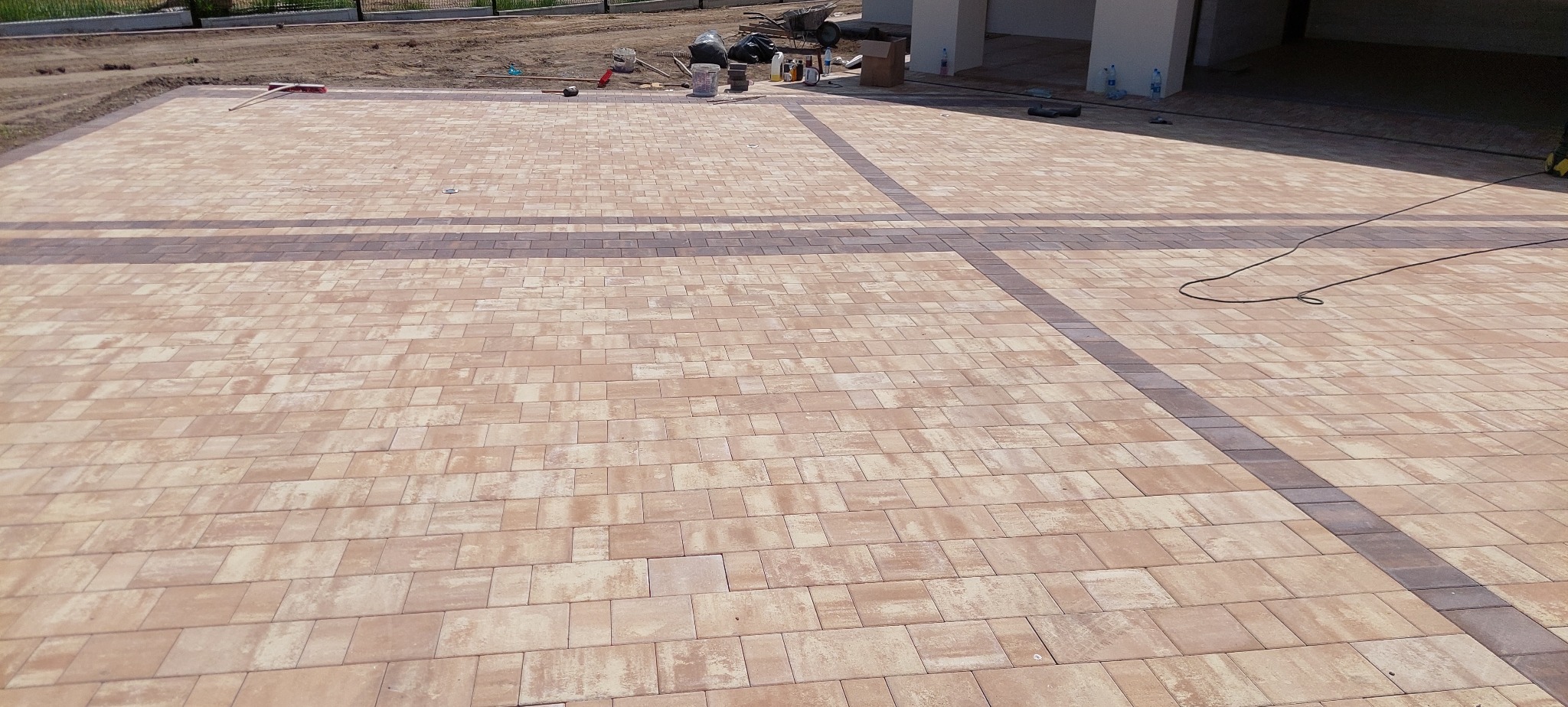 Ułożona kostka brukowa w beżowych odcieniach z ciemniejszymi pasami tworzącymi wzór krzyża. Widoczne narzędzia i materiały budowlane w tle. Całość sprawia wrażenie solidnego wykonania.