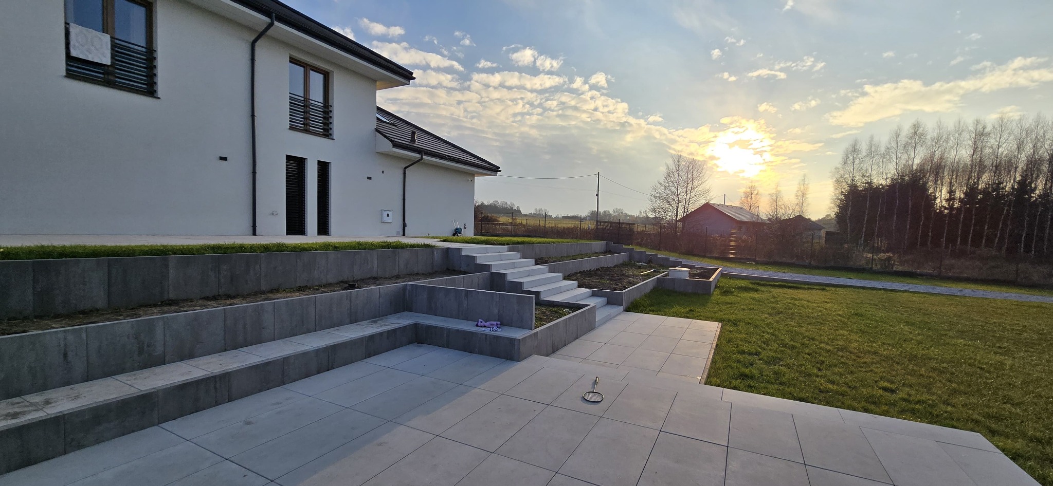 Nowoczesny taras z szarej kostki, schody z betonu architektonicznego prowadzące do ogrodu, biały dom w tle, zachód słońca.