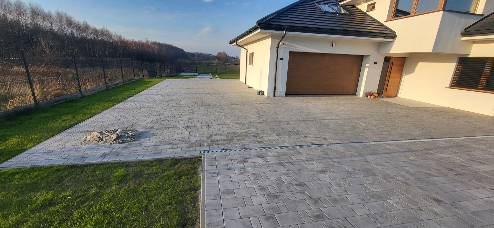 Szara kostka brukowa ułożona na podjeździe nowoczesnego domu z brązową bramą garażową i elementami zieleni. Widoczny fragment trawnika i ogrodzenie.