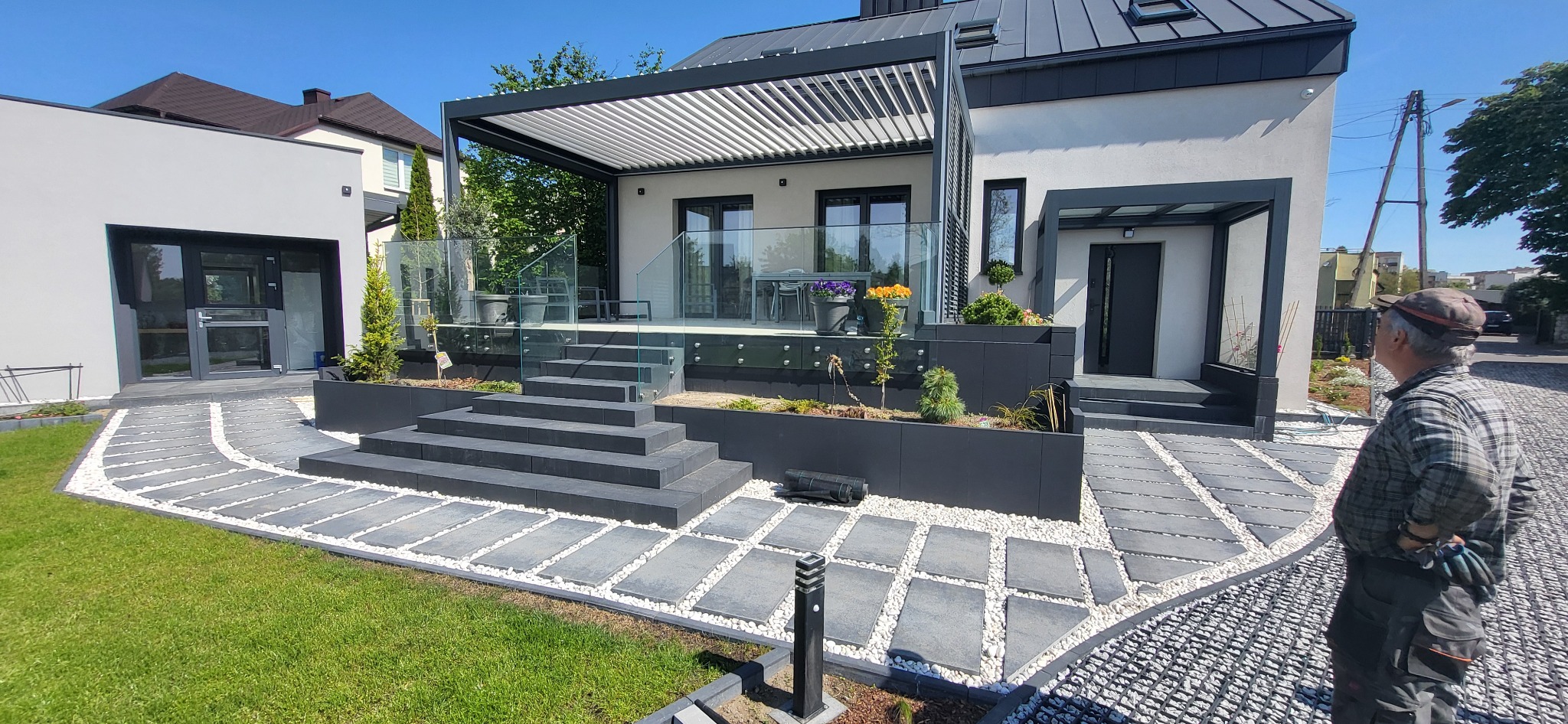 Nowoczesny ogród z tarasem i pergolą. Szara kostka brukowa ułożona w ścieżkę, białe kamienie ozdobne. Minimalistyczny design z elementami zieleni i szklanymi balustradami.