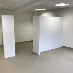 Wnętrze biurowe z dwiema ściankami działowymi z białych płyt, surowe wykończenie, widoczne łączenia płyt, jasna podłoga z płytek.