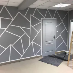 Ściana pomalowana na szaro z geometrycznym wzorem wykonanym białą taśmą malarską, drewniana drabina oparta o ścianę, zabezpieczona podłoga, widoczna puszka farby.