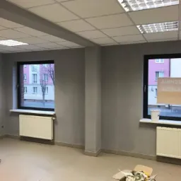 Wnętrze pomieszczenia po gładziowaniu ścian, widoczne dwa okna z grzejnikami, puste wiadro po farbie i karton na podłodze.