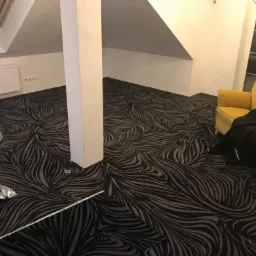 Pokój na poddaszu z nowo położoną, wzorzystą wykładziną dywanową w odcieniach szarości i czerni, widoczny fragment żółtej sofy i rolki wykładziny.