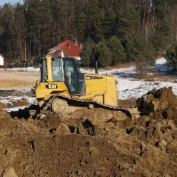 Żółty spychacz CAT D6N na nierównym, błotnistym terenie z resztkami śniegu, w tle las i domy.