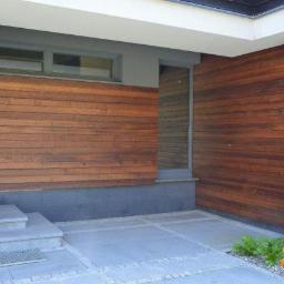 Bud-Ma - Nowoczesna elewacja domu z drewnianą okładziną poziomą, szara podmurówka, kamienne schody wejściowe i geometryczna nawierzchnia z kostki brukowej oraz mały klomb z żółtymi kwiatami.