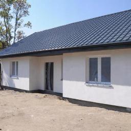 Bud-Ma - Nowo wybudowany, parterowy dom z ciemnoszarą, metalową dachówką, białymi ścianami i oknami zabezpieczonymi folią, widok z zewnątrz na tle drzew i nieba.