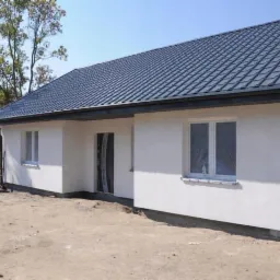 Nowo wybudowany, parterowy dom z ciemnoszarą, metalową dachówką, białymi ścianami i oknami zabezpieczonymi folią, widok z zewnątrz na tle drzew i nieba.