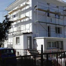 Bud-Ma - Pracownik na rusztowaniu maluje elewację dwupiętrowego domu w jasnym kolorze, widok zza metalowego ogrodzenia.