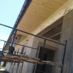 Bud-Ma - Budowa domu: widok z rusztowania na drewniany strop, betonową kolumnę i fragment ściany z pustaków. Na zewnątrz widać osobę i zieleń.