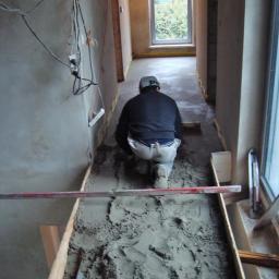 Bud-Ma - Pracownik w kasku wyrównuje wylewkę betonową w wąskim korytarzu podczas remontu, widoczne przewody elektryczne na ścianie i okno w oddali.